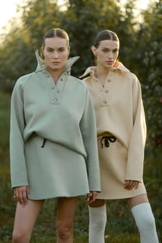 Two models in stylish tracksuits pose in apple orchard at sunset . The model is wearing a light-colored tracksuit and skirt.