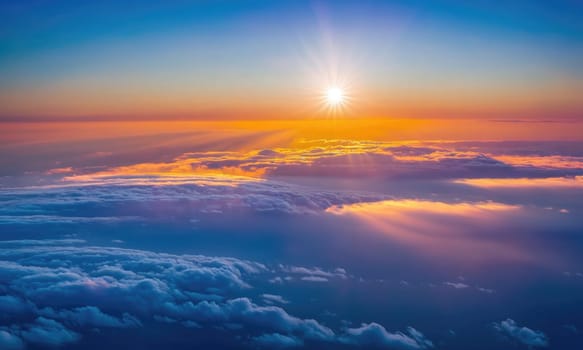 An aerial view of the setting sun casting light through the clouds. The sky is a vibrant orange, yellow, and blue.