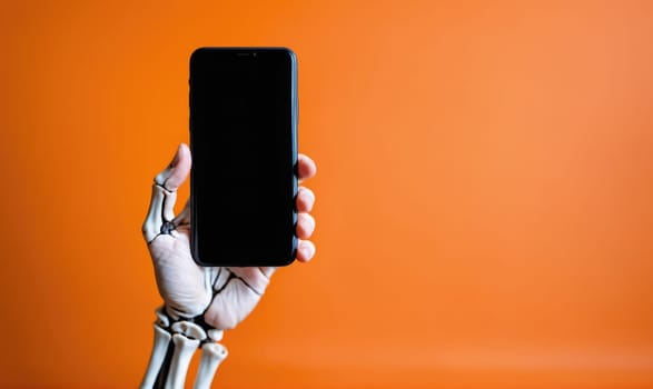 A skeletal hand holds a black smartphone vertically against an orange background. The hand is bare and the fingers are spread. The screen of the phone is black.