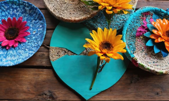 A sunflower stem is placed in a teal heart-shaped dish on a rustic wooden table. Several other decorative dishes, made from repurposed materials, are surrounding the sunflower.