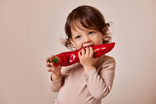 happy child girl bites fresh red pepper on beige background. healthy baby food. kid loves vegetables
