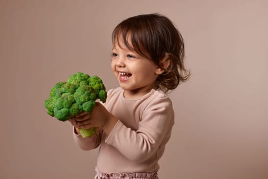 smiling child girl showing fresh green raw broccoli on beige background. healthy baby food. kid loves vegetables