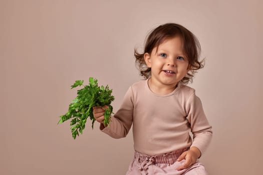happy child girl holding fresh green parsley on beige background. healthy baby food.