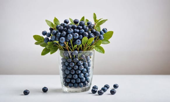 A glass vase filled with blueberries and green leaves is on a white table. Several blueberries are scattered on the table in front of the vase.