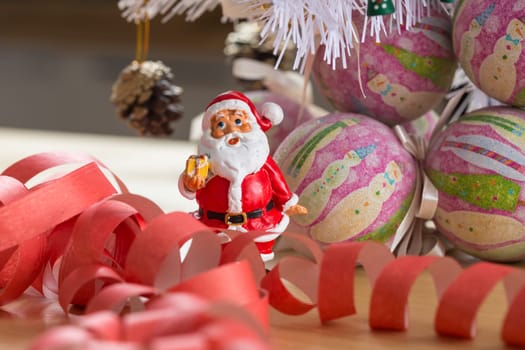 A small figurine of Santa Claus in a red and white suit holding a green sack, set against a blurred background. This festive ornament captures the spirit of Christmas.