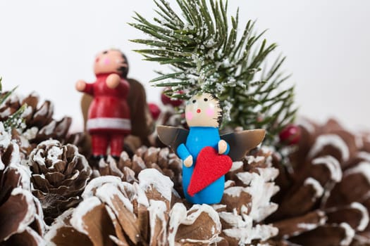 Christmas ornament featuring a small wooden angel holding a red heart, set against a festive background of pinecones and snow-dusted fir branches
