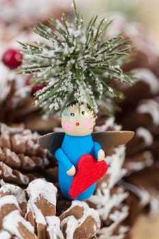 Christmas ornament featuring a small wooden angel holding a red heart, set against a festive background of pinecones and snow-dusted fir branches