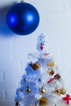 Small white Christmas tree decorated with golden and red ornaments, pine cones, and miniature Santa figurines on a wooden table against a white brick wall.