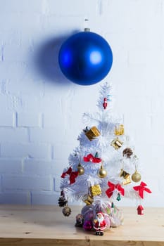 Small white Christmas tree decorated with golden and red ornaments, pine cones, and miniature Santa figurines on a wooden table against a white brick wall.