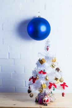 Small white Christmas tree decorated with golden and red ornaments, pine cones, and miniature Santa figurines on a wooden table against a white brick wall.
