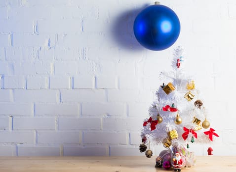 Small white Christmas tree decorated with golden and red ornaments, pine cones, and miniature Santa figurines on a wooden table against a white brick wall.