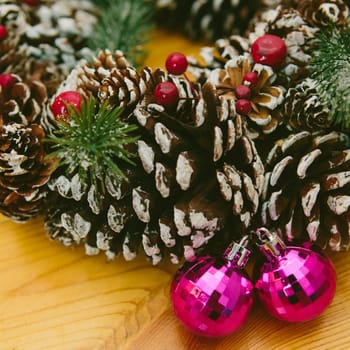 Holiday wreath adorned with snow-dusted pine cones, evergreen branches, red berries, and two shiny pink Christmas ornaments