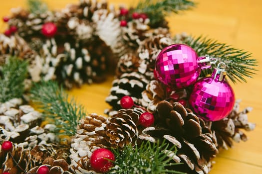 Holiday wreath adorned with snow-dusted pine cones, evergreen branches, red berries, and two shiny pink Christmas ornaments