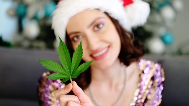A cheerful woman in a Santa hat holds a vibrant green cannabis leaf among colorful holiday decorations