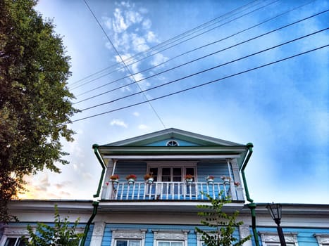 The blue wooden house features a balcony with flower pots, surrounded by trees under a clear sky at sunset.