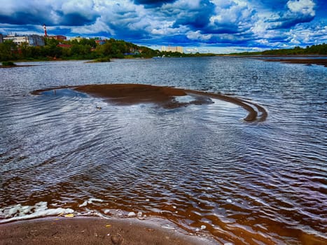 The river displays a calm surface with soft ripples, surrounded by lush greenery and distant buildings under a vibrant, cloudy sky.