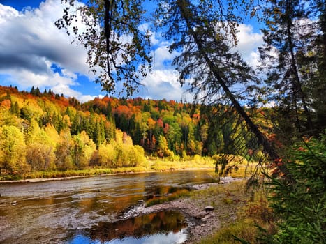 Vibrant autumn foliage surrounds a calm river, reflecting the colorful leaves under a bright sky with scattered clouds.