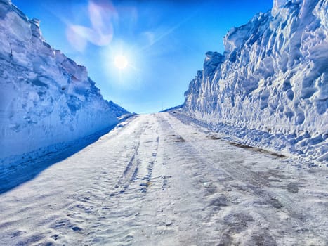 A deserted road flanked by high snowbanks shines under the vibrant sun, showcasing a tranquil winter day in a snowy landscape.