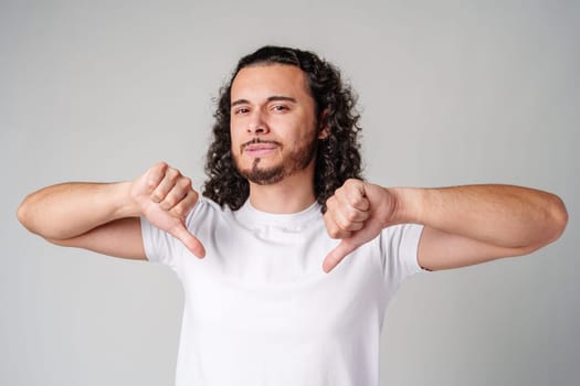 A young man with curly hair joyfully pointing down while wearing a plain white t-shirt against a neutral background close up