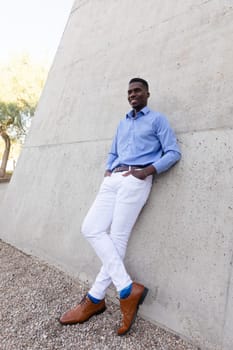 Full Size Handsome Stylish African American Man wearing White Jeans and Blue Shirt, Brown Belt, Shoes Outdoor, Concrete Wall on Background. Fashionable Middle Age Male in His 40s, Ivorian. Vertical.