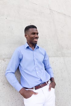 Portrait Of Handsome Smiling With Teeth African American Man wearing White Jeans and Blue Shirt, Brown Belt Outdoor, Concrete Wall on Background. Fashionable Male, Ivorian Vertical Plane