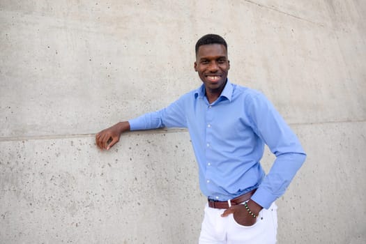 Portrait Of Handsome Smiling With Teeth African American Man wearing White Jeans and Blue Shirt, Brown Belt Outdoor, Concrete Wall on Background. Copy Space Fashionable Male, Ivorian. Horizontal.