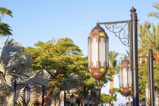 An elegant outdoor lantern with ornate details and colorful stained glass panels is featured, framed by tropical greenery under a clear blue sky. The decorative lighting fixture contrasts with the natural backdrop.
