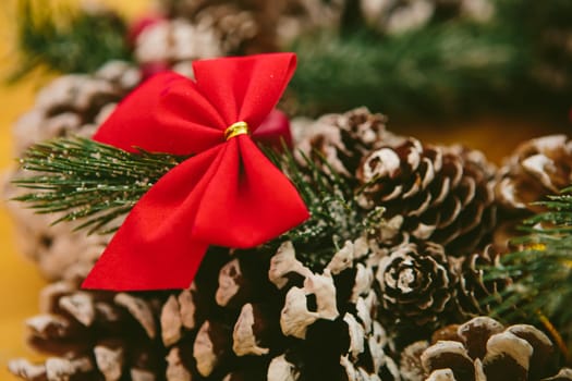 Festive red bow tied to a pine cone, part of a Christmas wreath. The vibrant red bow, accented with a gold ring, contrasts beautifully against the natural textures of pine cones and evergreen branches, creating a warm and joyful holiday atmosphere.