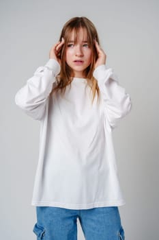 Young girl in a white sweater holding her head in discomfort against a minimalist background close up