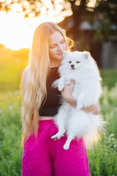 Young blonde woman is holding her white pomeranian dog in her arms while standing in a field at sunset