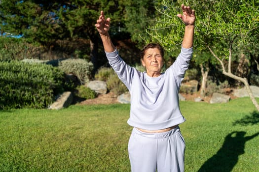 Determined senior woman in gray sportswear stretching arms in sunny park. Her focus reflects her commitment to staying active and healthy on bright sunny day