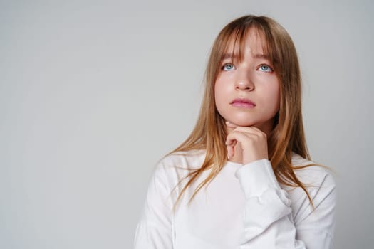Young woman in a white long-sleeve shirt thoughtfully poses with a hand on her chin in a minimalistic indoor setting