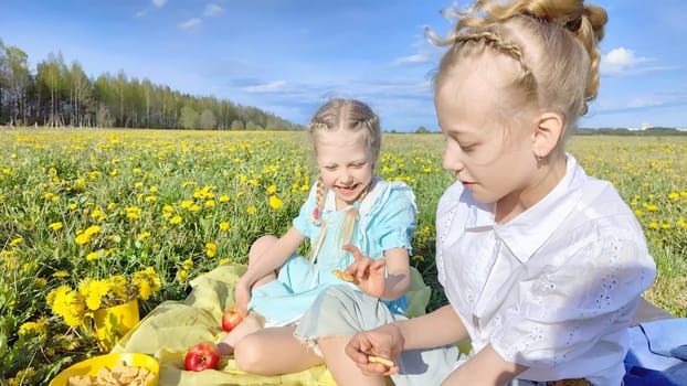 Two young girls laugh and share snacks in a lush green field adorned with blooming flowers under a clear blue sky.