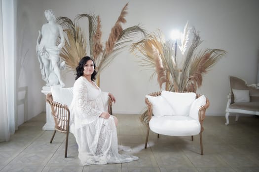 A plump woman poses gracefully in a delicate dress, surrounded by elegant decor in a softly lit indoor setting.