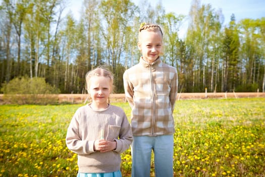 Two girls pose happily among blooming flowers, enjoying a warm spring day in a vibrant green landscape with trees in the background.