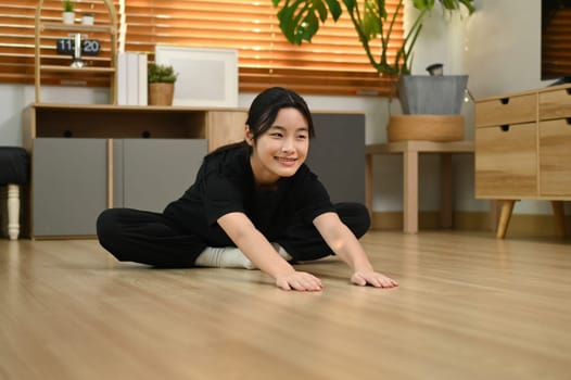 Pretty asian girl stretching on wooden floor getting ready for workout at home.