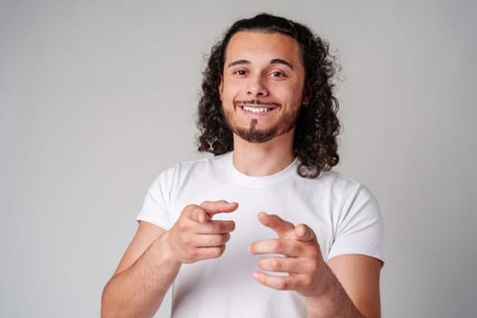 A smiling young man with curly hair playfully pointing directly at the viewer in a light-colored setting close up