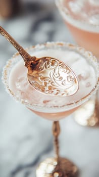 Pink cocktail in a coupe glass with a sugared rim is being stirred with an ornate gold spoon