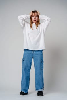 A teen girl in a loose white sweater looks surprised with her hands on her head against a plain gray backdrop close up