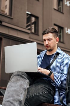 Young man working on a laptop while seated on a bench outside close up