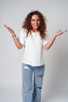 Cheerful woman with curly hair poses with open arms in a white shirt against a plain background close up