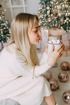 Blonde in a bright Christmas interior with a cup of hot drink with marshmallows. New Year's interior in pink tones. The winter holidays period.