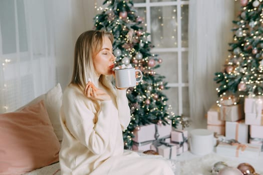 Blonde in a bright Christmas interior with a cup of hot drink with marshmallows. New Year's interior in pink tones. The winter holidays period.