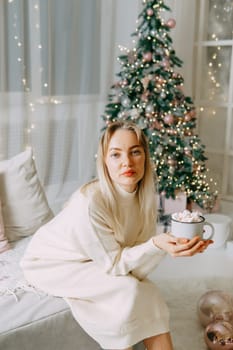 Blonde in a bright Christmas interior with a cup of hot drink with marshmallows. New Year's interior in pink tones. The winter holidays period.