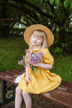A little girl in a yellow dress and straw hat wearing a bouquet of lilacs. A walk in a spring park, blossoming lilacs