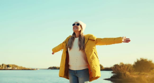 Happy smiling woman raising her hands up on sunny beach, enjoying amazing landscape, warm weather, wear hat, yellow jacket, standing on sea coast, nature background