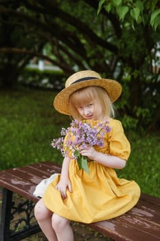 A little girl in a yellow dress and straw hat wearing a bouquet of lilacs. A walk in a spring park, blossoming lilacs