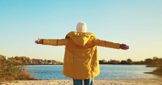 Back view of happy woman raising her hands up on sunny beach, enjoying amazing landscape, warm weather, wear hat, yellow jacket, standing on sea coast, nature background