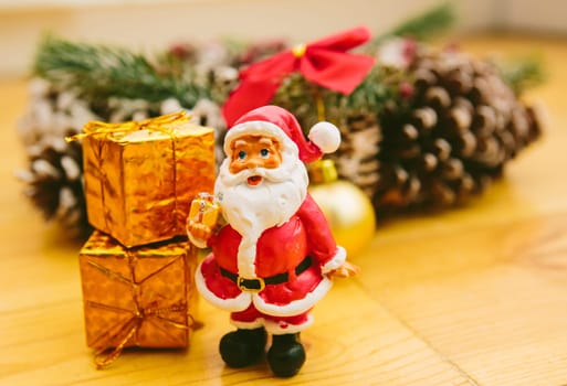 Christmas wreath decorated with pine cones, red bows, and evergreen branches sits on a wooden surface. In front of the wreath, a small Santa Claus figurine and golden ornaments add a cheerful holiday touch