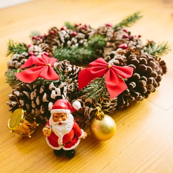 Christmas wreath decorated with pine cones, red bows, and evergreen branches sits on a wooden surface. In front of the wreath, a small Santa Claus figurine and golden ornaments add a cheerful holiday touch